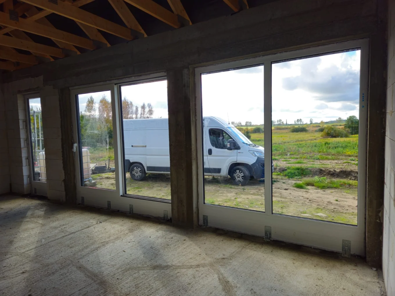 Wnętrze nowego budynku z zamontowanymi oknami wielkoformatowymi na parterze, za nimi widoczny biały dostawczy van i pejzaż