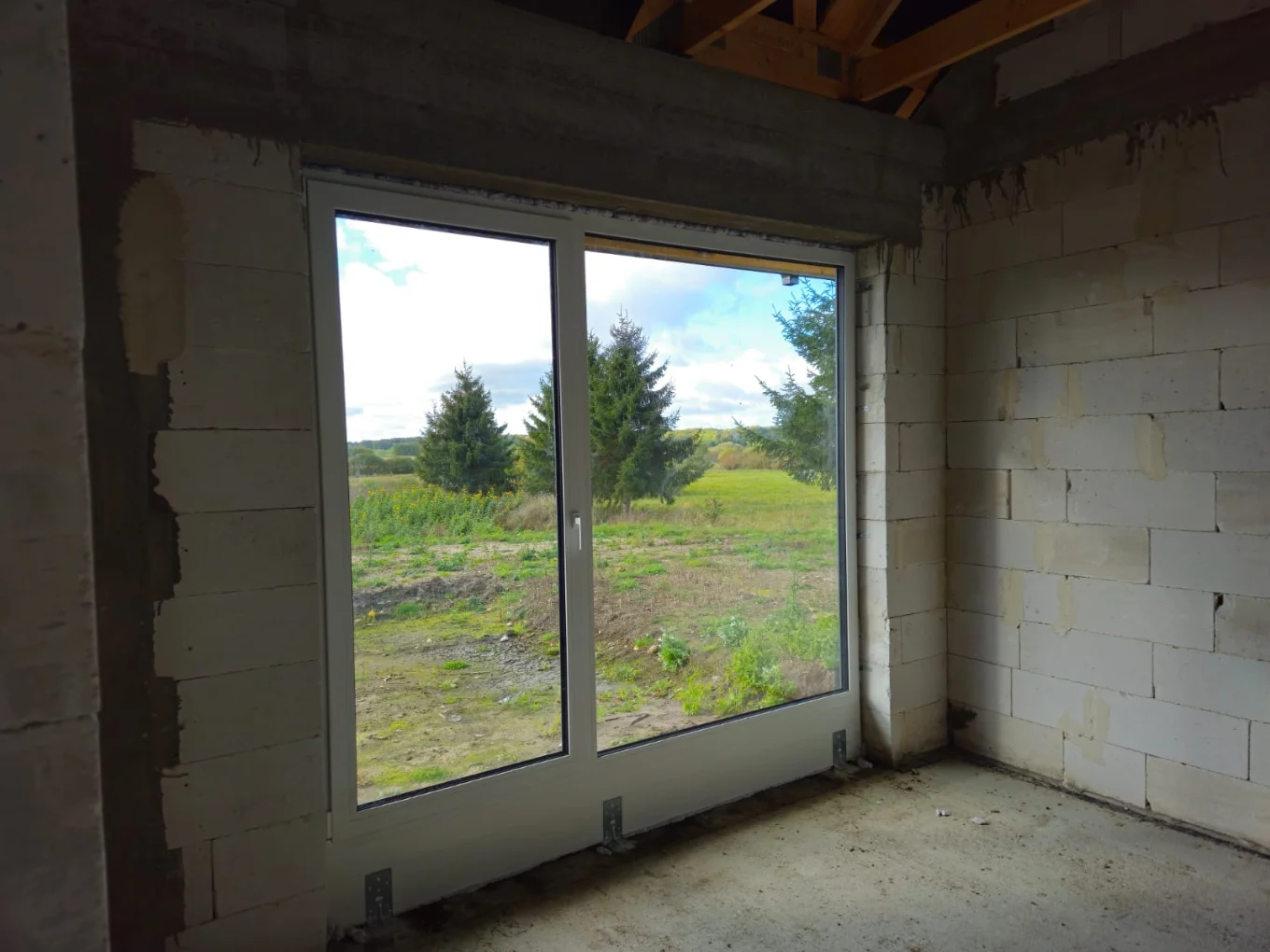 Surowe wnętrze budynku z zamontowanymi oknami tarasowymi i widokiem na zieleń