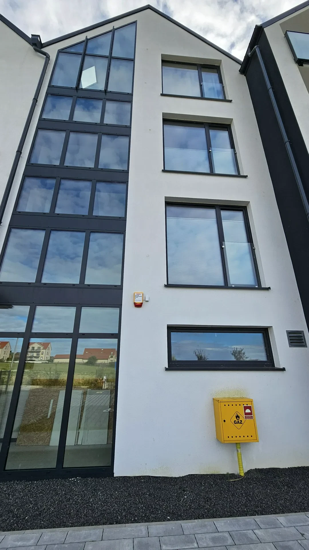 Biała fasada budynku z czarną przeszkloną klatką schodową i czterema oknami wielkoformatowymi