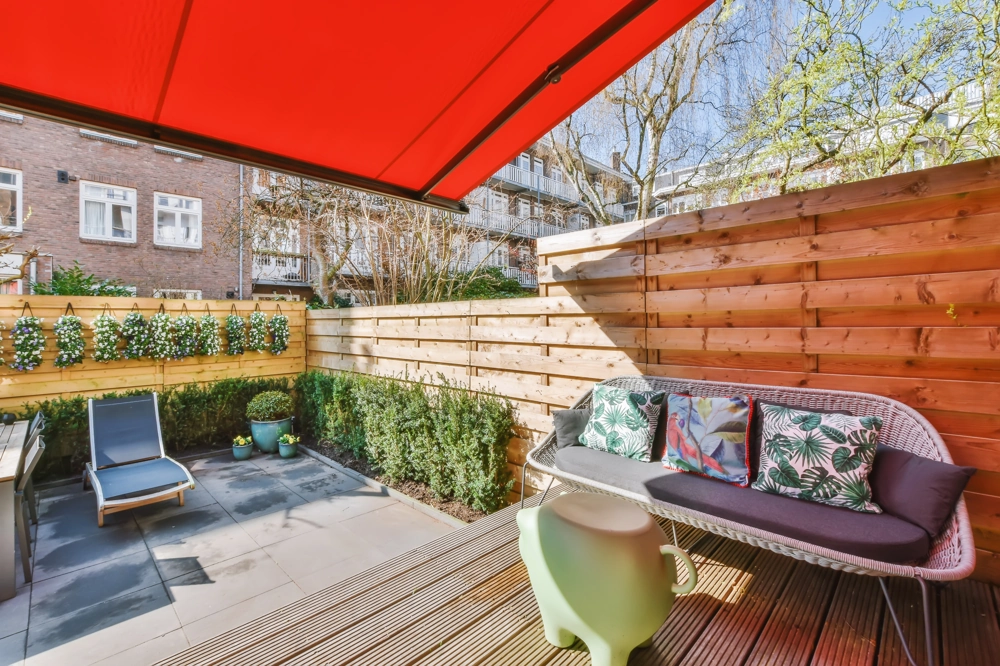 Czerwona markiza balkonowa nad tarasem z drewnianą zabudową i meblami ogrodowymi