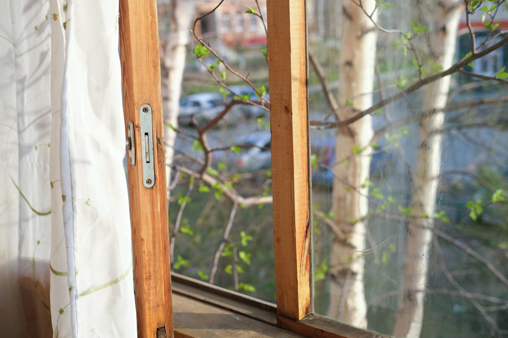 Otwarta drewniana rama okienna z białą zasłoną i widokiem na pąki na gałęziach – Montaż okien