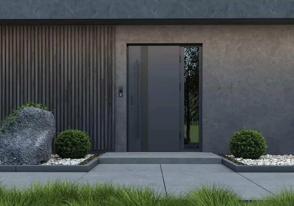 Szare drzwi wejściowe z bocznym naświetlem, betonowy podjazd i ozdobne krzewy przy elewacji