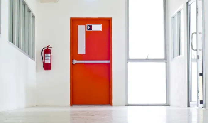 Czerwone drzwi ppoż z zamkiem antypanicznym i gaśnicą na białym korytarzu